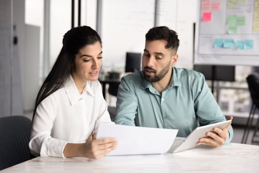Two coworkers at an office table review a printed document and a tablet while discussing benefits paperwork.