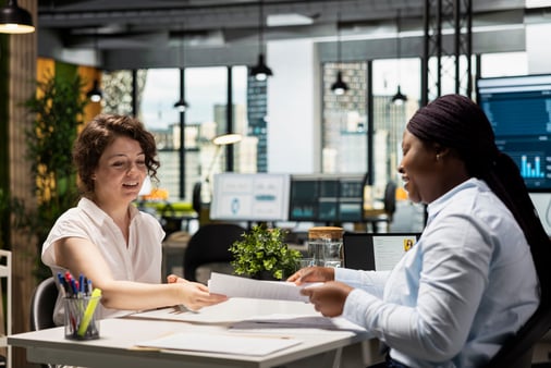 Two smiling coworkers at a desk in a bright, modern office pass paperwork between them, with a city skyline visible through the windows behind them.
