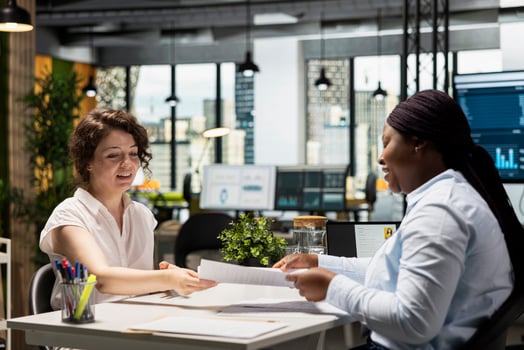 Two smiling coworkers at a desk in a bright, modern office pass paperwork between them, with a city skyline visible through the windows behind them.