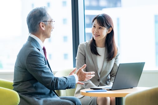 Two professionals in business suits talk across a table with a laptop in a sunlit office.