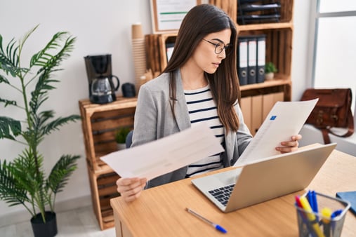 Woman in a gray blazer comparing two benefits documents at her desk during open enrollment.