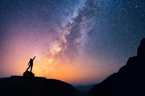 Silhouette of a person in front of a starry night sky, reaching upwards towards the stars. 