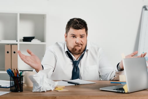 Man sitting at a desk with a laptop and shrugging 