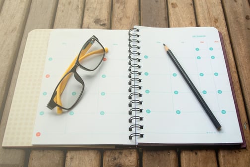 A planner that is open, with a pencil and a pair of eyeglasses sitting on top of it